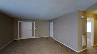 This single-floor residential space offers a straightforward and functional layout designed for comfortable everyday living. The main living area is finished with wall-to-wall beige carpeting that adds warmth underfoot, while the walls are painted in a soft, neutral taupe tone that creates a calm and understated ambiance throughout. The ceiling features a classic textured popcorn finish, adding subtle dimension overhead. A paneled front entry door with an adjacent sidelight panel provides natural light near the entryway, offering a welcoming point of arrival. White trim and baseboards run consistently throughout the space, providing clean contrast against the neutral wall color. An interior door leads to an additional room or hallway, suggesting a connected layout with multiple functional areas. A wall-mounted thermostat is visible, indicating individual climate control within the unit. The space benefits from a simple, open feel with no visual obstructions, making it highly adaptable for a variety of furniture arrangements and living configurations. Overall, the space presents a clean, move-in ready environment with classic residential finishes suitable for practical, day-to-day use.