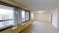 This single-floor space presents a spacious, open-plan layout finished in a clean, neutral palette. The room is carpeted throughout in a warm beige tone, contributing to a calm and understated ambiance. Large sliding windows along one wall allow generous amounts of natural light to flood the interior, framed by vertical blinds that offer privacy and light control. The windows appear to overlook an exterior balcony, suggesting elevated views of the surrounding area. A second window on the adjacent wall further enhances the brightness of the space. Baseboard heating units are positioned beneath the windows, providing efficient climate control integrated into the room's perimeter. The white ceiling is smooth and complemented by two classic pendant light fixtures, which provide overhead illumination and add a simple, timeless design element. The overall layout is unobstructed and versatile, lending itself well to a variety of furniture arrangements. The neutral tones of the walls, ceiling, and flooring create a cohesive and move-in-ready environment suited to a range of interior styles.