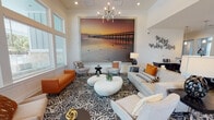 This thoughtfully designed single-floor clubhouse and leasing center offers a refined, coastal-inspired environment with an open, airy layout. The main lounge is an immediate highlight, featuring soaring ceilings, oversized windows that flood the space with natural light, and a dramatic large-scale coastal sunset mural serving as a commanding focal point. An elegant chandelier anchors the room, which is furnished with a curated mix of neutral and warm-toned seating arranged around a sculptural white coffee table, all set atop a bold geometric-patterned area rug. Decorative metallic wall art adds an organic, nature-inspired touch throughout the space. Adjacent to the lounge, a dedicated conference room provides a professional setting with a substantial dark wood table, leather armchairs, a wall-mounted display screen, and generous window banks that invite abundant daylight. The kitchenette area features light wood-style flooring, dark cabinetry, a granite-topped breakfast bar with rattan barstools, stainless steel appliances, and a ceiling-mounted monitor. The reception area includes a granite-topped service counter, pendant lighting, and a well-organized corridor leading to additional rooms. Throughout, light wood flooring, high ceilings, and consistent coastal-modern design elements create a cohesive and welcoming atmosphere.