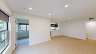 This single-floor residence offers a clean, contemporary design throughout, characterized by crisp white walls, smooth ceilings, and light-toned hardwood flooring that flows seamlessly from room to room. The open-concept living and dining area is bright and airy, benefiting from natural light and providing a clear sightline toward the kitchen. A doorway leads to an outdoor balcony, extending the living space and offering views of the surrounding exterior. The kitchen is a functional highlight of the space, featuring rich dark cabinetry that creates a striking contrast against white quartz countertops. Stainless steel appliances — including a gas range, refrigerator, built-in microwave, and dishwasher — are integrated throughout, complemented by a wall-mounted range hood. A dedicated sink with a window above adds both utility and natural light to the cooking area. The bedrooms are generously proportioned with clean white walls and the same consistent hardwood flooring. The primary bedroom includes a built-in closet system with frosted glass sliding doors, offering ample and organized storage. Additional rooms provide flexible use and are well-suited for various configurations. The overall design aesthetic is minimal and modern, creating a cohesive and functional living environment.