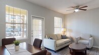 Located at 927 VFW Blvd in San Antonio, Texas, this single-floor unit offers a thoughtfully designed layout that balances comfort and functionality. The open-concept living and dining area serves as the heart of the space, finished with durable wood-style flooring and neutral gray walls that create a clean, modern aesthetic. Multiple windows dressed with slatted blinds flood the area with natural light, while a ceiling fan overhead adds to the comfort of the room. The living area features a cozy seating arrangement, and the adjacent dining space comfortably accommodates a full dining table and chairs, making it ideal for everyday living and entertaining. A door from the living area provides direct outdoor access. The kitchen is well-equipped with a two-tone cabinetry design — dark lower cabinets contrasting with crisp white upper cabinets — complemented by light quartz-style countertops and black appliances, including a full-size refrigerator and a gas range with overhead ventilation. The bedroom is a serene retreat, bathed in natural light from a large window with outdoor views of surrounding greenery. The bathroom features a shower-tub combination and connects to a dedicated walk-in closet, offering practical storage solutions throughout.