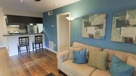 This single-floor space showcases a well-designed open layout that seamlessly connects the living area to the kitchen. The living room is anchored by a bold teal accent wall, which adds a vibrant pop of color and serves as a striking visual focal point. A beige sofa dressed with coordinating blue and green throw pillows sits comfortably against this wall, complemented by a tall arc floor lamp that provides warm ambient lighting. A contemporary abstract canvas artwork adorns the teal wall, enhancing the room's modern aesthetic. The flooring throughout is warm-toned hardwood, adding texture and a sense of continuity across the space. A doorway framed in crisp white trim leads further into the unit, contributing to the open, airy feel. The kitchen is visible from the living area and features a raised breakfast bar with bar seating, stainless steel appliances, and dark countertops — ideal for casual dining or entertaining. Overhead ventilation registers are integrated into the walls, ensuring functional climate control. The overall ambiance is modern and inviting, with a cohesive color palette that balances neutral furnishings against the bold teal walls.