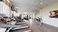 Located at 7405 Long Lake Rd in Columbus, Ohio, this fitness center offers a spacious, single-floor layout designed to accommodate a full range of workout activities. The open floor plan is finished with durable speckled rubber flooring throughout, providing both functionality and easy maintenance.

The cardio zone is generously stocked with multiple treadmills and elliptical machines, each equipped with individual digital displays for tracking performance. A selection of stationary bikes and a cable weight machine round out the strength training options, while a dedicated free-weight and bench press area adds further versatility to the workout space.

Large windows line multiple walls, flooding the interior with natural light and creating an open, airy atmosphere. Ceiling fans are strategically positioned across the ceiling to ensure comfortable airflow during workouts. Recessed lighting supplements the natural illumination for evening hours. Wall-mounted flat-screen televisions are installed near the cardio equipment, keeping members entertained during their sessions.

Motivational fitness artwork decorates the neutral-toned walls, contributing to an energizing ambiance. A mirrored wall section enhances the sense of spaciousness. The facility also includes an adjacent restroom accessible through glass-paned interior doors, adding convenience for users. Multiple entry and exit points ensure easy access throughout the space.