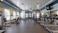 Located at 1010 Versant Dr in Brandon, Florida, this well-appointed fitness center offers a comprehensive workout facility on a single floor. The space is defined by its distinctive tray ceiling accented with deep teal and soft blue tones, framed by elegant crown molding and recessed lighting that creates a bright, energizing atmosphere. A ceiling fan adds comfort to the workout environment.

The facility is generously equipped for a full range of exercise routines. The cardio zone features multiple treadmills and elliptical machines arranged along a mirrored wall, which visually expands the space and allows users to monitor their form. A wall-mounted television is positioned above the mirrors for entertainment during workouts. The strength training area includes a multi-station cable machine, various selectorized weight machines, adjustable benches, and a well-stocked dumbbell rack. Medicine balls and stability balls are neatly organized on a dedicated rack.

Durable rubber flooring covers the entire workout area, providing safety and sound absorption. Multiple windows along the walls allow natural light to fill the room while offering views of the surrounding outdoor greenery. Entry is through glass-paneled doors, contributing to an open, welcoming feel. Towel stations and a water dispenser are conveniently available for users. Overall, the space delivers a functional, clean, and comfortable fitness environment.