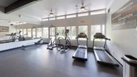 This fitness center is a thoughtfully designed, single-floor facility that offers a comprehensive workout environment for all fitness levels. The space is bathed in natural light, courtesy of a generous band of clerestory windows that run along the upper walls, offering views of the surrounding greenery and creating an open, energizing atmosphere. High ceilings with ceiling fans enhance ventilation and comfort throughout.

The cardio zone is well-appointed with multiple treadmills, elliptical trainers, a stationary bike, and a rowing machine, all positioned to take advantage of the natural light and outdoor views. Full-length mirrors along one wall expand the sense of space and allow users to monitor their form during workouts.

A dedicated strength and functional training area features a full dumbbell rack, kettlebells, medicine balls, TRX suspension trainers, battle ropes, and a multi-functional cable and pull-up rig. This zone is distinguished by bold gray and orange accent walls, adding a dynamic, motivating energy to the space.

An adjoining room, accessible through glass-paned sliding doors, provides additional space for stretching, yoga, or small group exercise. Dark rubber flooring throughout ensures durability and safety. A motivational typography mural adorns one wall, reinforcing the facility's focus on wellness and determination. Overall, the fitness center is a functional, modern, and inviting space designed to support a wide range of fitness goals.
