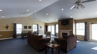 This single-floor space is designed to provide a comfortable and functional gathering environment. The main room features soaring vaulted ceilings with recessed lighting and dual ceiling fans, creating an airy, open atmosphere. The layout seamlessly integrates a lounge area and a dining space within the same open floor plan, making the room feel expansive and versatile.

The lounge area is anchored by a pair of plush, dark brown sofas arranged around a wooden coffee table, offering a relaxed setting for conversation or leisure. A wall-mounted flat-screen television is positioned above a classic white-mantled fireplace, serving as a natural focal point on one side of the room. On the opposite end, a dining table with multiple chairs provides ample seating for group meals or activities.

Natural light flows generously through multiple windows fitted with decorative curtains and a set of glass-paneled doors, which also offer access to the outdoors. The warm beige and neutral-toned walls are accented by a two-tone paint treatment with a chair-rail detail, adding a touch of architectural refinement. Soft carpeting throughout contributes to the welcoming, comfortable character of the space, while a small accent chair offers an additional cozy nook near the windows.