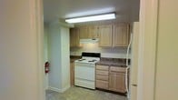 Located in Silver Spring, Maryland, this single-floor space offers a practical and straightforward layout. The kitchen is a standout feature, designed with light natural wood cabinetry that lines the upper and lower walls, providing ample storage. Dark granite-style countertops offer a subtle contrast to the lighter tones of the cabinetry, creating a balanced aesthetic. White appliances, including an electric range and a full-sized refrigerator, complement the clean, neutral color palette. An overhead range hood is centrally positioned above the stove, adding functionality to the cooking area. Recessed fluorescent lighting illuminates the kitchen efficiently, ensuring a well-lit workspace. The flooring consists of neutral-toned tiles, contributing to the overall understated and practical character of the space. A fire extinguisher is conveniently mounted near the kitchen entrance, reflecting attention to safety. The overall ambiance is clean and utilitarian, making it a functional living space well-suited for everyday use in the Silver Spring area.