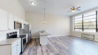 This single-floor unit offers a clean, modern design highlighted by wood-look flooring that flows throughout the entire space. The open-concept layout seamlessly connects the kitchen, dining, and living areas, creating a bright and functional environment. Large grid-pane windows flood the living area with natural light and offer outdoor views, while a ceiling fan provides year-round comfort.

The kitchen is well-appointed with tall white shaker-style cabinetry extending toward the ceiling, a classic white subway tile backsplash, neutral quartz-style countertops, and a full suite of stainless steel appliances including a French door refrigerator with ice maker, electric range, and over-the-range microwave. A central kitchen island with a built-in sink and pendant lighting above serves as both a prep space and casual dining area.

The bedroom features high ceilings, neutral walls, and a spacious reach-in closet with double doors and a clothes rod. An additional closet near the entryway provides extra storage with shelving. The bathroom is finished with a white vanity, a subway tile tub-surround, chrome fixtures, and wood-look flooring. A dedicated laundry room houses a full-size top-loading washer and dryer alongside the water heater, adding everyday convenience to this thoughtfully designed unit.