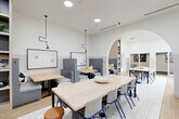 Located on South University Avenue in Ann Arbor, this thoughtfully designed communal space offers a versatile, single-floor layout ideal for collaboration, focused work, and social interaction. The interior is characterized by crisp white walls with a two-tone wainscoting effect, warm wood-tone flooring, and a mix of patterned tile and hardwood surfaces that define distinct zones throughout.

The main gathering area features a large communal table with upholstered chairs and round stools in a mix of blue and tan, anchored by pendant lighting with adjustable arm fixtures overhead. A distinctive arched passageway connects this central zone to an adjacent area, adding an architectural focal point that lends a sense of openness and flow.

Private booth-style seating lines one wall, offering semi-enclosed spaces with gray upholstered benches and individual tables — ideal for small group meetings or focused work. Built-in open shelving with decorative accents and greenery adds warmth to the transition between zones.

A quieter alcove accessed through the arch contains individual study desks paired with high-backed wooden chairs, wall-mounted sconce lighting, and a calm lavender-gray lower wall treatment. Curated artwork punctuates the walls throughout, enhancing the space's contemporary aesthetic. Recessed ceiling lighting ensures consistent, bright illumination across all areas.