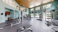 This single-floor fitness center is thoughtfully designed to accommodate a wide range of exercise needs. The main gym area is the centerpiece of the facility, boasting soaring double-height ceilings clad in warm wood paneling, aqua-toned shiplap walls, and an abundance of natural light streaming through clerestory windows positioned along the upper perimeter. The space is outfitted with an extensive array of strength and cardio equipment, including weight machines, free weights, dumbbells, medicine balls, stability balls, cable systems, treadmills, ellipticals, a rowing machine, and a functional training rig. Full-length mirrors line the walls, enhancing the sense of space and allowing users to monitor their form. Pendant lighting and wall-mounted televisions add to the modern, energetic atmosphere. Adjacent to the main gym is a dedicated indoor cycling studio featuring a row of stationary spin bikes, wood flooring, mirrored walls, mounted televisions, and recessed lighting. A separate multi-purpose studio space with hardwood floors and large windows overlooking an outdoor pool area provides an ideal setting for yoga, stretching, or group fitness classes. The facility's cohesive design and comprehensive equipment selection make it a fully functional destination for all fitness levels.