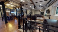 This one-of-a-kind clubhouse is housed within a thoughtfully converted historic mill, blending industrial heritage with modern comfort throughout its expansive single-floor layout. The space retains numerous original architectural elements, including exposed wooden ceiling beams, black steel structural columns, and dramatic large-scale mill wheels mounted as permanent decorative features — each serving as a striking reminder of the building's storied past.

Warm, wide-plank hardwood floors run throughout, complementing the textured plaster walls and recessed lighting that illuminate the space. Black wrought iron railings border an open pit area at the heart of the clubhouse, where original millworks machinery remains visible below floor level, offering an authentic industrial focal point.

Multiple seating areas are arranged across the open layout, each furnished with dark leather sofas and area rugs, creating inviting spaces for gathering. A wall-mounted television and natural light from several windows enhance the lounge atmosphere. Exposed ductwork and piping contribute to the industrial aesthetic without compromising comfort.

The kitchen area is well-appointed with rich wood cabinetry, granite countertops, stainless steel appliances including a refrigerator, microwave, and sink — ideal for entertaining and community events.