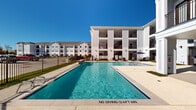 Located at 10742 Freehill Street in Houston, TX, this outdoor amenity area is part of a modern multi-story residential community finished in crisp white siding. The centerpiece of the space is a large rectangular swimming pool with clear, blue water, surrounded by a broad concrete pool deck that provides ample room for lounging. Depth markings and stainless steel entry rails reflect a focus on safety and accessibility. The pool deck is enclosed by a sleek black metal fence, offering both security and open sightlines to the surrounding landscape under expansive Texas skies.

Adjacent to the pool, a covered outdoor living area serves as a shaded retreat, equipped with ceiling fans to promote airflow and comfort. This covered patio features a built-in countertop with a sink, functioning as an outdoor kitchen prep station. Rattan-style lounge chairs with blue cushions are arranged to encourage relaxed gatherings. Large sliding glass doors connect this covered space to an interior area, enhancing the indoor-outdoor flow.

The overall ambiance is clean, contemporary, and well-maintained, with lush green grass accents framing the hardscape. The multi-story residential buildings visible in the background provide architectural context, completing a polished community setting designed for outdoor enjoyment.