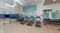 This fitness and recreation facility is thoughtfully designed to support a wide range of athletic and wellness activities. The space is anchored by a generously sized multi-use court with a polished hardwood floor, marked for both racquetball and basketball. The court is enclosed by full glass walls and doors, allowing clear sightlines into the playing area while maintaining an open, airy feel. Padded blue wall panels line the lower portion of the court, providing both protection and a bold visual accent against the bright white upper walls. Recessed ceiling lighting illuminates the court evenly throughout.

Adjacent to the court, the fitness area is outfitted with a comprehensive selection of equipment, including multiple treadmills, an elliptical trainer, a free weight station with a full rack of dumbbells, and additional strength and conditioning machines. Light wood-style flooring runs throughout the gym area, complementing the clean white walls accented with teal paint and decorative botanical artwork. Full-length mirrors along one wall expand the sense of space and support proper form during workouts.

The entry vestibule, finished in teal, features a water cooler and informational postings, providing a functional transition into the facility. The overall design is modern and well-maintained, creating an inviting environment for both casual exercisers and serious athletes.