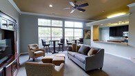 This single-floor communal space is thoughtfully designed to offer a comfortable and inviting atmosphere for both casual gatherings and quiet relaxation. The main lounge area is anchored by a generous gray sofa accented with earth-toned throw pillows, complemented by patterned armchairs and upholstered ottomans that create a warm, layered seating arrangement. A ceiling fan with integrated lighting overhead adds a functional yet classic touch to the space.

Large, multi-pane windows line one wall, flooding the interior with abundant natural light and offering pleasant views of lush greenery outside. Beneath the windows, a bistro-style high-top table with swivel bar chairs provides an additional seating zone, perfect for informal conversations or individual use.

The space transitions seamlessly into an adjacent area distinguished by a bold yellow accent wall and a pass-through counter or service window, suggesting a kitchenette or refreshment station nearby. Neutral gray walls and carpeted flooring throughout create a cohesive, calming backdrop that ties the various seating zones together. Recessed lighting supplements the natural light, ensuring the space remains bright and welcoming at any hour. Overall, the layout balances openness with a sense of coziness, making it a versatile and functional communal environment.