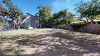This community amenity space includes two distinct areas designed to support an active lifestyle. The fitness center is a well-appointed, single-level facility finished with patterned carpet flooring and a bold color scheme of orange and blue accent bands that run along the upper walls. Large windows along one side of the room flood the space with natural light while offering views of an outdoor pool area, creating an open and energizing atmosphere. The gym is comprehensively equipped with multiple treadmills, an elliptical trainer, upright and recumbent stationary bikes, a rowing machine, a multi-function cable/strength machine, and a full rack of dumbbells arranged in front of a large wall-mounted mirror. Recessed lighting and a ceiling fan ensure the space remains bright and comfortable during workouts. A dedicated entry door with exterior access provides convenient, direct entry to the facility. The outdoor dog park is a generously sized, open-air ground-level area enclosed by a sturdy wood privacy fence. The grounds are shaded by a canopy of mature deciduous trees, offering a natural and comfortable environment. The park sits adjacent to a multi-story brick building and includes a water station for pets, making it a practical and pleasant space for dogs and their owners to enjoy the outdoors.