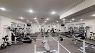 This fitness center is a fully outfitted exercise facility designed to accommodate a comprehensive range of workout routines. The space is anchored by a generous array of cardio equipment, including multiple treadmills and elliptical machines, all arranged to provide ample room for movement between stations. A dedicated strength training area features free weights, barbells, and weight benches, offering options for both beginner and experienced lifters. Wall-to-wall mirrors line the perimeter, creating a sense of expanded space while allowing users to monitor their form during exercise. The ceiling is fitted with recessed lighting that illuminates the entire floor evenly, contributing to a clean, functional atmosphere. Flat-screen televisions are mounted at strategic points around the room, providing entertainment during workouts. The flooring consists of durable carpet suited for a fitness environment, and ventilation is supported by visible ceiling vents. Windows along one wall allow for natural light to supplement the interior lighting. The overall design is practical and utilitarian, prioritizing functionality and ease of use throughout the single-level layout.