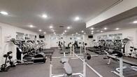 This fitness center is a fully outfitted exercise facility designed to accommodate a comprehensive range of workout routines. The space is anchored by a generous array of cardio equipment, including multiple treadmills and elliptical machines, all arranged to provide ample room for movement between stations. A dedicated strength training area features free weights, barbells, and weight benches, offering options for both beginner and experienced lifters. Wall-to-wall mirrors line the perimeter, creating a sense of expanded space while allowing users to monitor their form during exercise. The ceiling is fitted with recessed lighting that illuminates the entire floor evenly, contributing to a clean, functional atmosphere. Flat-screen televisions are mounted at strategic points around the room, providing entertainment during workouts. The flooring consists of durable carpet suited for a fitness environment, and ventilation is supported by visible ceiling vents. Windows along one wall allow for natural light to supplement the interior lighting. The overall design is practical and utilitarian, prioritizing functionality and ease of use throughout the single-level layout.