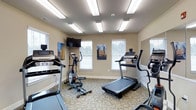 Located on State Street, this single-floor fitness room offers a practical and well-organized space for cardiovascular exercise. The room is outfitted with several cardio machines, including treadmills, an elliptical cross-trainer, and additional aerobic equipment, providing a variety of workout options. A wall-mounted flat-screen television is positioned for easy viewing during exercise sessions, adding convenience for users during longer workouts. Natural light filters into the room through multiple windows fitted with white blinds, creating a bright and welcoming atmosphere. The overhead lighting, consisting of several flush-mounted fluorescent panels, ensures consistent illumination throughout the space. The floor is covered in a patterned, low-pile carpet suited for gym use. Neutral-toned walls contribute to the clean, functional aesthetic of the room. A large mirror is mounted along one wall, enhancing the sense of openness within the space. Decorative artwork displayed on the walls adds a subtle visual element to the otherwise utilitarian environment. Overall, the fitness room presents a clean, well-lit, and functional setting designed to support an active lifestyle.