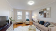 Montclair Gardens, Unit A5 is a well-appointed single-floor residence that blends classic architectural details with modern updates throughout. Warm hardwood floors run continuously across the living spaces, complementing the crisp white walls and elegant crown molding that frame each room.

The living room is a welcoming, light-filled space anchored by multiple double-hung windows that invite abundant natural light. Classic cast-iron radiators add a charming period touch, while the generous layout comfortably accommodates a full seating arrangement.

The kitchen has been thoughtfully updated with sleek, high-gloss white cabinetry, grey quartz countertops, a white tile backsplash, and a full suite of stainless steel appliances, including a gas range, microwave, dishwasher, and refrigerator. The hardwood flooring continues seamlessly into this space.

The bedroom is a serene retreat featuring multiple windows on two walls, filling the room with natural light and offering views of the surrounding neighborhood. Ample wall space and hardwood floors make it both functional and inviting.

The bathroom is finished in crisp white with marble-style tile flooring, a tub-shower combination, and a window for natural ventilation. Overall, Unit A5 offers a cohesive and comfortable living environment with timeless details throughout.