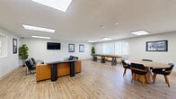 Located at 923 Maplewood Dr in Cedar Falls, Iowa, this well-appointed single-floor space offers a generously open layout designed for both leisure and socializing. Light wood-look flooring runs throughout, creating a warm, cohesive aesthetic that complements the clean white walls and textured ceilings.

The main living area is thoughtfully divided into distinct functional zones. A comfortable lounge area features a tan leather sectional sofa, sleek black accent chairs, and a wall-mounted flat-screen TV, all anchored by a stylish area rug and a dark sculptural side table. Recessed panel lighting overhead ensures the space is evenly and brightly lit.

Adjacent to the lounge, a round wooden dining table with matching black chairs provides a casual gathering spot, bathed in natural light from multiple windows fitted with vertical blinds that frame outdoor views of the surrounding greenery. A full-length shuffleboard table serves as a standout recreational feature, offering an engaging activity for residents or guests.

The entryway welcomes visitors with a clean, minimalist aesthetic — double glass-panel interior doors, a white console table, and flush-panel ceiling lights set a polished tone. Framed black-and-white photography adorns the walls throughout, adding a curated, community-oriented character to the overall space.