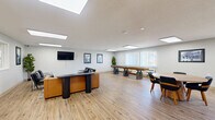 Located at 923 Maplewood Dr in Cedar Falls, Iowa, this well-appointed single-floor space offers a generously open layout designed for both leisure and socializing. Light wood-look flooring runs throughout, creating a warm, cohesive aesthetic that complements the clean white walls and textured ceilings.

The main living area is thoughtfully divided into distinct functional zones. A comfortable lounge area features a tan leather sectional sofa, sleek black accent chairs, and a wall-mounted flat-screen TV, all anchored by a stylish area rug and a dark sculptural side table. Recessed panel lighting overhead ensures the space is evenly and brightly lit.

Adjacent to the lounge, a round wooden dining table with matching black chairs provides a casual gathering spot, bathed in natural light from multiple windows fitted with vertical blinds that frame outdoor views of the surrounding greenery. A full-length shuffleboard table serves as a standout recreational feature, offering an engaging activity for residents or guests.

The entryway welcomes visitors with a clean, minimalist aesthetic — double glass-panel interior doors, a white console table, and flush-panel ceiling lights set a polished tone. Framed black-and-white photography adorns the walls throughout, adding a curated, community-oriented character to the overall space.