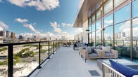 This sophisticated rooftop terrace offers a striking urban retreat designed for both relaxation and entertaining. The expansive open-air deck wraps around the building's upper level, bordered by sleek black metal railings that frame unobstructed panoramic views of the surrounding city skyline, greenery, and neighboring architecture.

The terrace is thoughtfully divided into multiple distinct zones. One area features generously sized lounge seating with woven rattan frames, plush cushions in neutral tones, and navy blue accent pillows arranged around a central fire feature — ideal for casual gatherings. A separate section provides alfresco dining with dark-framed tables paired with woven chairs, positioned to take full advantage of the elevated cityscape.

A dramatic floor-to-ceiling glass curtain wall runs along one side of the terrace, flooding adjacent interior spaces with natural light while maintaining a seamless visual connection between indoors and out. The smooth concrete flooring extends throughout, providing a clean, contemporary aesthetic that complements the building's modern architectural language.

The terrace is oriented to capture abundant natural light and open sky, with cloud-filled blue skies and city landmarks visible from virtually every vantage point. The overall ambiance is refined yet approachable, blending high-end outdoor furnishings with an architectural design that prioritizes views, openness, and urban connection.