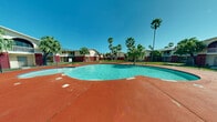This outdoor amenity space centers around a generously sized freeform swimming pool with clear turquoise water, creating a resort-like atmosphere ideal for relaxation and recreation. The pool deck is finished in a distinctive red-painted concrete surface that extends broadly around the pool's organic, curved shape, providing ample space for lounge seating and poolside activities. A dedicated shaded pergola structure is positioned along one side of the deck, offering a covered retreat from the sun.

Surrounding the pool area, decorative white gravel landscaping beds add visual contrast against the red deck, while well-maintained green lawns border the perimeter. Tall, mature palm trees are distributed throughout the space, lending a tropical character and providing natural shade. The entire pool area is enclosed by a chain-link security fence, ensuring a safe and defined recreational zone.

The backdrop consists of two-story residential buildings with a bold beige and red exterior color scheme, featuring balconies and arched architectural detailing. The open-air setting benefits from abundant natural light and clear, expansive sky views, reinforcing the warm, sunny climate of the location. Overall, the space presents a well-appointed communal outdoor environment with a strong tropical aesthetic.