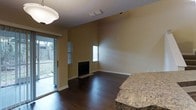 This two-story property features an open and airy living area defined by notably high ceilings that amplify the sense of volume throughout the main level. Dark hardwood flooring runs seamlessly across the space, providing a rich contrast against the warm, neutral-toned walls finished with a textured ceiling overhead.

A centrally positioned fireplace serves as a focal point along one wall, framed by clean, simple surroundings that keep the aesthetic understated and functional. Natural light filters into the room through multiple windows fitted with shuttered blinds, as well as a full sliding glass door that opens directly to an outdoor area, offering views of a fenced backyard with greenery visible beyond.

A granite-topped surface — likely a kitchen counter or bar — is partially visible at the edge of the frame, suggesting an open-concept layout that connects the living area to an adjacent kitchen space. A ceiling-mounted light fixture provides additional illumination to complement the natural light. The overall ambiance is clean and functional, with a neutral palette and quality finishes that create a comfortable, versatile living environment across both floors.