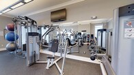 Located at 199 Cedar Park Boulevard in Northampton County, Pennsylvania, this fitness center offers a dedicated, single-floor space designed to support a full range of exercise routines. The gym is outfitted with a comprehensive selection of strength training equipment, including multi-function cable machines and resistance stations from recognized brands such as Life Fitness, providing users with versatile options for targeted muscle training. Cardio equipment is also present, rounding out the facility's offerings for a well-balanced workout environment. Full-length mirrors line the walls, visually expanding the space and allowing users to monitor their form during exercise. The flooring features durable, textured rubber matting well-suited for heavy equipment and high-traffic use. Crown molding along the upper walls adds a refined architectural touch, lending the space a polished appearance. A wall-mounted flat-screen television is positioned for easy viewing during workouts, enhancing the overall experience. Natural light filters in through windows, contributing to a bright and energizing atmosphere. The space is thoughtfully laid out to accommodate multiple users simultaneously while maintaining an open, comfortable feel throughout.