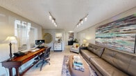 Located at 1025 S Crater Rd in Petersburg, Virginia, this single-floor unit offers a thoughtfully organized layout across three distinct areas. The combined kitchen and living room serves as the heart of the space, where light wood-style flooring, track lighting, and a glass-paneled entry door create a bright and welcoming atmosphere. The kitchen features white cabinetry, dark countertops, and stainless steel appliances, including a refrigerator and range, all efficiently arranged in a galley-style configuration. The open living area accommodates a comfortable seating arrangement highlighted by abstract wall art and lush indoor greenery. The dining room provides a dedicated workspace and meeting area, furnished with a large wooden table, upholstered chairs, and metal filing cabinets, complemented by a textured ceiling and a window that brings in natural light. Urban-themed artwork adorns the walls, adding a contemporary touch. The bathroom is finished with light wood-look tile flooring, a white vanity with cabinet storage, a mirrored medicine cabinet, and clean, neutral-toned walls. The exterior of the building features classic gray siding and is surrounded by mature trees and landscaped grounds, offering a pleasant street-side presence.