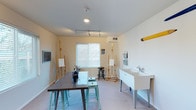 This single-floor art studio is thoughtfully designed to provide a functional and inspiring workspace for artists. The room features neutral white walls and light-colored flooring, creating a clean, distraction-free environment that allows artwork to take center stage. A large window fitted with horizontal blinds allows natural light to fill the space, complementing the adjustable track lighting mounted on the ceiling, which ensures consistent, directed illumination across multiple work areas.

Several wooden easels are positioned throughout the room, accommodating multiple artists simultaneously. A sturdy, dark-topped worktable occupies the center of the studio, surrounded by teal metal stools that provide casual, flexible seating. Art supplies, including brushes and pencils, are organized and readily accessible on the table surface.

A deep utility sink with a faucet is installed along one wall, offering convenient cleanup for wet media work. A wall-mounted pegboard or organizer nearby provides additional storage for tools and materials. A small chalkboard on the opposing wall serves as a functional teaching or planning surface. The overall ambiance is bright, orderly, and purposefully arranged to support creative work.