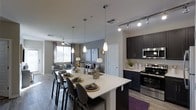 This single-floor residence offers a thoughtfully designed open-concept layout that seamlessly connects the kitchen, dining, and living areas into one cohesive and functional space. The kitchen is a standout feature, boasting dark espresso cabinetry that contrasts elegantly with a bright white island countertop, stainless steel appliances, and a mosaic tile backsplash. Pendant lighting hangs above the expansive island, which doubles as a breakfast bar with seating for multiple guests. Track lighting overhead adds to the bright, well-lit atmosphere. The dining area flows naturally from the kitchen, centered around the island and framed by modern bar-style seating. The open living room beyond is anchored by a ceiling fan, neutral-toned furnishings, and large windows that invite natural light deep into the space. Warm hardwood-style flooring runs throughout, adding continuity and a sense of warmth. Neutral gray walls with subtle accent tones create a contemporary backdrop throughout. The overall ambiance is modern, clean, and inviting, making this space well-suited for both relaxed daily living and casual entertaining.