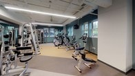 Located at 310 W 15th St in Minneapolis, this single-floor fitness center offers a comprehensive workout facility designed to support a wide range of exercise needs. The space is organized into distinct functional zones connected by a bright, welcoming corridor finished with light wood-look tile flooring, warm walnut accent wall panels, and curated artwork that lends a polished, modern aesthetic.

The main exercise area is generously sized, featuring an extensive lineup of cardio equipment including treadmills, ellipticals, and stationary bikes, each equipped with individual monitors for entertainment or workout tracking. Overhead, linear LED lighting illuminates the space efficiently, while windows along the walls allow natural light to filter in and provide views of the surrounding neighborhood greenery.

A dedicated strength training zone offers a robust selection of cable machines, weight racks, and resistance equipment, catering to both beginners and experienced athletes. The flooring transitions between cushioned rubber matting in workout zones and smooth surfaces in transitional areas, providing both safety and comfort.

A separate multipurpose room features mirrored walls, open floor space, and exercise balls, making it ideal for stretching, yoga, or group fitness. A hydration station with dual water fountains, set against a striking blue tile accent wall, is conveniently positioned near the corridor. The facility's teal accent walls, exposed utility infrastructure, and thoughtful layout create a functional yet visually engaging environment.
