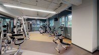Located at 310 W 15th St in Minneapolis, this single-floor fitness center offers a comprehensive workout facility designed to support a wide range of exercise needs. The space is organized into distinct functional zones connected by a bright, welcoming corridor finished with light wood-look tile flooring, warm walnut accent wall panels, and curated artwork that lends a polished, modern aesthetic.

The main exercise area is generously sized, featuring an extensive lineup of cardio equipment including treadmills, ellipticals, and stationary bikes, each equipped with individual monitors for entertainment or workout tracking. Overhead, linear LED lighting illuminates the space efficiently, while windows along the walls allow natural light to filter in and provide views of the surrounding neighborhood greenery.

A dedicated strength training zone offers a robust selection of cable machines, weight racks, and resistance equipment, catering to both beginners and experienced athletes. The flooring transitions between cushioned rubber matting in workout zones and smooth surfaces in transitional areas, providing both safety and comfort.

A separate multipurpose room features mirrored walls, open floor space, and exercise balls, making it ideal for stretching, yoga, or group fitness. A hydration station with dual water fountains, set against a striking blue tile accent wall, is conveniently positioned near the corridor. The facility's teal accent walls, exposed utility infrastructure, and thoughtful layout create a functional yet visually engaging environment.