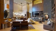 This single-floor space presents an open, contemporary lobby lounge designed with both comfort and style in mind. The most striking architectural feature is the dramatic double-height ceiling, which lends a grand sense of openness and airiness to the entire area. Recessed lighting throughout the ceiling is complemented by a bold, industrial-style rectangular pendant chandelier that serves as a central design focal point.

The space is divided into distinct functional zones. A casual seating area features plush, dark brown upholstered chairs and sofas arranged around a low coffee table atop a patterned area rug, ideal for relaxed conversation or leisure. Adjacent to this, white modern lounge chairs offer a contrasting, lighter seating option. In the background, a well-appointed bar or café counter with bar stools provides a social dining area.

A floor-to-ceiling built-in shelving unit lines one wall, housing books, decorative items, and a mounted flat-screen television, adding both function and visual warmth. The warm neutral color palette — incorporating beige, taupe, and rich brown tones — combined with light hardwood flooring creates a cohesive, inviting ambiance throughout the space.