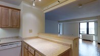 This single-story space is characterized by warm hardwood flooring that extends throughout the majority of the layout, lending a cohesive and inviting feel to every room. The floor plan, as seen from above, reveals several generously sized rooms arranged in an open, interconnected configuration, offering a practical and comfortable flow between spaces.

The kitchen is a notable feature, equipped with light wood cabinetry, laminate countertops, and a distinctive pass-through window that opens directly into an adjacent living area. This pass-through design enhances connectivity between the kitchen and the rest of the space while allowing natural light to travel between rooms. Track lighting in the kitchen provides focused, functional illumination.

A full bathroom is centrally located within the layout, featuring standard fixtures including a toilet and what appears to be a combined tub and shower unit. Its central placement ensures convenient access from multiple areas of the space.

The remaining rooms are spacious and versatile, with layout proportions that suggest potential use as living, dining, or sleeping areas. Natural light enters through windows visible in several rooms, brightening the warm wood-toned interiors and contributing to an open, airy ambiance throughout the single-level floor plan.