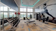 Located at 13645 E Colonial Dr in Orlando, FL, this contemporary fitness center is designed to deliver a premium workout experience in a spacious, single-floor layout. The facility immediately impresses with its soaring double-height ceilings accented by teal LED cove lighting, creating a vibrant and energizing atmosphere throughout.

Large, floor-to-ceiling windows line the walls, inviting generous amounts of natural light and providing open views of the surrounding outdoor greenery. The flooring features a stylish patchwork carpet in neutral tones with blue accent blocks, adding a modern aesthetic that carries throughout the space.

The strength training area is thoroughly equipped with a full rack of dumbbells, a multi-functional cable machine, a Smith machine with weight storage, adjustable benches, medicine balls, and a heavy punching bag suspended from a TRX-style rig. A bold motivational mural reading "The Sport of Fitness Has Arrived" anchors the free weights section alongside a wall of mirrors.

The cardio zone offers a lineup of Life Fitness treadmills, stationary bikes, and elliptical machines, all positioned to face the expansive windows. Ceiling fans provide additional airflow comfort. The clean white walls, recessed lighting, and thoughtfully organized equipment layout make this facility both functional and visually impressive.