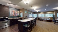 This single-floor community amenity space is thoughtfully designed to offer residents a variety of functional and social environments. The entrance foyer welcomes visitors with soaring ceilings, abundant natural light from transom windows above glass doors, and tasteful artwork lining the walls, setting an inviting tone throughout.

The central social lounge is a standout feature, anchored by a large white-topped kitchen island with bar seating and dark cabinetry. Decorative teal glass pendant lights hang overhead, complementing the bold accent wall. A full kitchenette with stainless steel appliances, including a refrigerator, microwave, and dishwasher, makes the space ideal for gatherings.

An adjoining seating area offers comfortable lounge furniture, including a leather sofa and upholstered chairs, bathed in natural light from bamboo-shaded windows. A cozy fireplace nook features a tile-surround gas fireplace, wall-mounted television, and dining tables — perfect for relaxed socializing.

The fitness center is vibrant and energetic, with bold teal and yellow accent walls, large windows overlooking the outdoors, and a comprehensive selection of cardio and strength training equipment including treadmills, ellipticals, and cable machines. Wide, carpeted corridors with warm crown molding trim connect all areas seamlessly, ensuring easy navigation throughout the facility.
