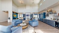 Located on Curtis Road in Champaign, Illinois, this single-story community clubhouse is designed to offer residents a well-rounded social and recreational hub. The centerpiece is a striking main lounge with soaring double-height ceilings, dramatic sputnik chandeliers, and rich teal accent walls adorned with decorative artwork. Built-in shelving flanks a fireplace with a mounted flat-screen TV, creating an inviting gathering space filled with plush blue sofas and armchairs arranged around a wood-and-stone coffee table. A full-service kitchen features dark cabinetry, a mosaic tile backsplash, stainless steel appliances, and a large quartz-topped island with woven barstool seating. Large windows in the kitchen overlook an outdoor pool area, flooding the space with natural light. A dedicated game and recreation room offers patterned carpeting, a billiards table, a large wall-mounted Scrabble board, a TV lounge, and ample open floor space for activities. An adjacent multipurpose room with hardwood-style flooring and wall-mounted displays provides flexible event space. Large sliding glass doors lead to an outdoor covered porch with rocking chairs, offering pleasant views of the surrounding landscape. Package lockers and additional amenity spaces round out this thoughtfully designed community facility.