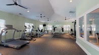 Located at 8439 Dorchester Rd in North Charleston, SC, this single-story fitness center offers a comprehensive and thoughtfully organized workout facility. The main exercise area is generously proportioned, featuring vaulted ceilings with multiple ceiling fans for optimal air circulation, recessed lighting, and abundant natural light streaming through multiple windows fitted with blinds. The space is finished with durable rubber flooring throughout, providing a safe and functional surface for a wide range of activities. Cardio equipment includes treadmills, elliptical trainers, a stationary bike, and a recumbent bike, all arranged to maximize floor space. A dedicated strength training zone houses a multi-station cable machine, weight benches, and plate-loaded machines, complemented by wall-mounted mirrors that enhance the sense of space. An interior window connects the main gym to a separate free weights room, which features warm hardwood-style flooring, sky-blue walls, recessed lighting, and its own natural light source. This room is stocked with a full dumbbell rack, kettlebells, medicine balls, and a stability ball. A connected studio space offers an inviting setting for yoga or personal training sessions, complete with a wall-mounted TV. The facility also includes a community bulletin board and clearly marked exits, reflecting a well-maintained, community-oriented environment.