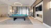 This contemporary amenity lounge in California offers a well-appointed, single-floor space designed for both recreation and relaxation. Light hardwood flooring flows throughout, anchoring the clean, modern aesthetic defined by crisp white walls and recessed lighting.

The recreation area centers around a regulation-size billiards table set atop a textured area rug, illuminated by a striking circular pendant light. Adjacent to it, a sophisticated lounge area features blue and neutral upholstered seating arranged around a central coffee table, anchored by a floor-to-ceiling marble-surround fireplace with a wall-mounted flat-screen TV above.

A full-length glass wall system with roller shades lines one side of the space, flooding the interior with natural light and offering views of a landscaped outdoor terrace. This seamless indoor-outdoor connection enhances the open, airy ambiance of the lounge.

A dedicated dining area accommodates multiple seating groups with white tables and modern chairs, positioned near a fully equipped kitchen featuring dark cabinetry, stainless steel appliances, and an island — ideal for catered events or casual gatherings. Wall sconces and cove lighting add warmth throughout. The space is thoughtfully designed to serve as a versatile, upscale communal amenity.