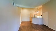 This single-floor residence offers a thoughtfully designed open-concept layout that connects the living area to the kitchen, creating a seamless and functional flow throughout the space. Rich, dark hardwood flooring extends across the main living area, providing a warm and inviting foundation that contrasts beautifully with the light, neutral-toned walls. The space features smooth ceilings with subtle architectural detailing, including a defined ceiling transition that adds visual interest and dimension to the interior. A ceiling fan is centrally positioned in the kitchen area, providing both practical airflow and a finishing touch to the room. The kitchen is equipped with light wood cabinetry and a raised breakfast bar counter, which serves as a natural dividing element between the cooking and living spaces while also offering casual seating. Stainless steel appliances, including a refrigerator and microwave, are visible and contribute to the kitchen's modern, functional character. The living area is generously proportioned, offering ample room for furnishings and everyday living. Recessed lighting supplements the overall brightness of the space. The combination of hardwood floors, open layout, and clean architectural lines gives this residence a comfortable, contemporary ambiance that is both practical and visually appealing.