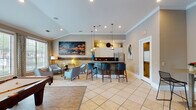Located at 13100 W Bellfort Ave in Houston, TX, this single-floor clubhouse facility offers a well-appointed communal space designed for comfort and social engagement. The interior is finished with large-format cream tile flooring throughout, crown molding, and recessed lighting that creates a bright, welcoming atmosphere.

The main gathering area serves as the centerpiece of the space, featuring a striking kitchen bar adorned with a mosaic stone surround and white upper cabinetry. Pendant lights hang above the bar counter, which is equipped with a stainless steel refrigerator and sink. Teal-upholstered bar stools provide comfortable seating at the counter, while nearby lounge chairs and curated artwork — including bold abstract canvases — lend an upscale residential feel.

Adjacent to the main lounge is a café-style seating area furnished with bistro-height tables and sleek wire-frame chairs. Decorative wall shelving and framed artwork accent the neutral-toned walls, contributing to the space's modern aesthetic.

A dedicated billiards room offers leisure with a full-size pool table set atop hardwood-style flooring, a wall-mounted television, and large windows that frame views of an outdoor pool area. The vaulted ceiling in this room adds an open, airy quality. Together, these areas create a versatile and visually cohesive community hub.