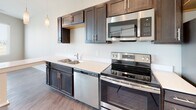 This thoughtfully designed single-floor residence offers a clean, modern aesthetic throughout, with a cohesive neutral color palette, wood-look hard surface flooring, and textured ceilings that add subtle character to each space.

The kitchen is a standout feature, showcasing rich dark-stained cabinetry with sleek brushed nickel hardware, light quartz countertops, and a full suite of stainless steel appliances including an electric range, over-the-range microwave, dishwasher, and refrigerator. A double-basin sink with a modern faucet anchors the counter space, while upper cabinetry provides generous storage. Pendant lighting accents the kitchen, enhancing its contemporary feel.

The open living area extends from the kitchen, bathed in natural light from large windows that frame outdoor views. The space feels airy and welcoming, with clean white walls and durable flooring underfoot transitioning to plush carpet in the bedroom area.

A dedicated laundry area accommodates a stacked GE washer and dryer unit, offering practical in-unit convenience. The hallway connects the living spaces efficiently, with dark cabinetry and a white countertop vanity visible nearby, suggesting a well-organized bathroom. Overall, this residence delivers a balance of style and functionality in a compact, single-level layout.