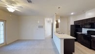This single-floor space is designed with an open-concept layout that seamlessly connects the kitchen and living area. The kitchen is a standout feature, equipped with dark espresso cabinetry, light granite countertops, and a peninsula that doubles as a breakfast bar with an integrated sink. Two pendant lights hang above the peninsula, providing focused task lighting while adding a modern aesthetic touch. A black refrigerator anchors the kitchen, complementing the dark cabinetry.

The living area extends from the kitchen, offering a generous, open feel underfoot with plush, neutral-toned carpeting. The white walls and standard-height ceilings with recessed lighting contribute to a clean, bright atmosphere throughout. A hallway visible from the main living space leads to additional rooms, suggesting a functional and well-organized floor plan. Ceiling-mounted HVAC vents are distributed throughout, ensuring climate control across the space. The overall design palette — neutral walls, dark kitchen fixtures, and light granite surfaces — creates a contemporary and cohesive aesthetic suitable for a variety of interior styles.