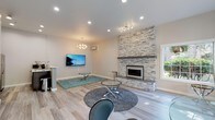 This thoughtfully designed single-floor space showcases a seamless open-concept layout that connects the living, dining, and kitchen areas with a cohesive modern aesthetic. Light-toned hardwood flooring flows throughout, lending warmth and continuity to the entire space.

The living area is anchored by a dramatic floor-to-ceiling stacked stone fireplace with a sleek linear electric insert and a solid mantel shelf, serving as a bold architectural focal point. Recessed lighting and a stylish chandelier complement the space, while large windows invite abundant natural light and views of lush outdoor greenery.

An expansive secondary living or flex area features a distinctive angled bay window with a built-in bench ledge and is accessible through elegant French doors, offering versatility for a variety of uses. This area is accented by contrasting warm taupe walls, adding depth and character.

The kitchen is efficiently designed with rich dark-toned cabinetry, stainless steel appliances, a mosaic tile backsplash, and a peninsula with a clean white countertop that provides both workspace and informal seating. A wall-mounted mini-split system ensures climate comfort throughout.

Overall, the space presents a polished, contemporary atmosphere with carefully curated design details that balance functionality and style.