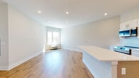 This well-appointed single-floor residence offers a clean, modern aesthetic throughout, defined by a neutral white-and-grey palette, recessed lighting, and warm hardwood-style flooring that flows seamlessly from room to room. The open-concept living and kitchen area forms the heart of the space, providing a generous, light-filled environment ideal for both everyday living and entertaining. Large windows invite natural light and offer views of the surrounding greenery, enhancing the airy ambiance of the main living area.

The kitchen is thoughtfully designed with crisp white shaker-style cabinetry reaching toward the ceiling, offering abundant storage. A full suite of stainless steel appliances — including a range, over-the-range microwave, dishwasher, and refrigerator — complement the bright white quartz countertops and a subtle mosaic tile backsplash. A substantial island with a clean white surface extends the kitchen into the living space, providing additional prep and seating opportunities.

The bathroom features a full tub-and-shower combination with a curved chrome rod, white tile flooring, and a sleek white vanity with modern hardware, maintaining the consistent contemporary style found throughout the unit. Louvered closet doors provide practical storage solutions. Overall, this space presents a polished, move-in-ready environment with refined finishes and a cohesive modern design.