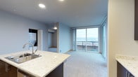 This well-appointed single-floor residence showcases a thoughtfully designed open-concept layout that seamlessly connects the kitchen, living, and dining areas. The kitchen features elegant light-toned granite or quartz countertops with a dark cabinetry base, a stainless steel sink, and recessed lighting that adds a clean, modern feel to the space. The living area is finished with plush wall-to-wall carpeting and soft blue-grey walls that create a calm, welcoming atmosphere. A full-width sliding glass door leads to a private balcony, flooding the interior with natural light and offering sweeping panoramic views of the water and surrounding cityscape. The balcony provides an ideal setting for enjoying the scenery throughout the day. Toward the back of the unit, a hallway leads to additional rooms, with a closet and a stacked washer and dryer unit conveniently accessible, adding a practical in-unit laundry feature. The overall design balances contemporary aesthetics with functional living, highlighted by recessed ceiling lights, neutral tones, and clean architectural lines. This space is well-suited for those seeking a modern, light-filled home with standout outdoor views.