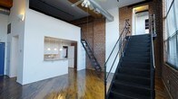 This two-story industrial loft showcases a striking blend of raw architectural character and contemporary design. The main living area commands attention with soaring double-height ceilings that amplify the sense of open space, while rich, wide-plank hardwood floors add warmth and depth throughout. Exposed brick walls run the full height of the interior, lending an authentic, urban aesthetic that anchors the space with texture and history. Overhead, visible ductwork and ceiling-mounted track lighting underscore the industrial design ethos.

A sleek kitchen is tucked behind a pass-through cutout in the wall, featuring pendant lighting above a clean white countertop bar — ideal for casual dining or entertaining. White cabinetry and appliances keep the kitchen bright and functional.

A bold black metal staircase with decorative cross-brace detailing rises prominently from the main floor, serving as both a functional element and a striking architectural focal point. It leads to the second level, offering a lofted upper floor that overlooks the main living area below.

Natural light filters in through windows, complementing the warmth of the hardwood and the texture of the brick. The overall ambiance is one of purposeful, well-preserved industrial character paired with the comfort of a modern living space.
