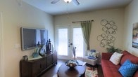Located in Grand Prairie, Texas, this single-floor residence offers a warm and welcoming atmosphere highlighted by thoughtful design details. The living area serves as the heart of the home, featuring rich hardwood-style flooring that grounds the space with a sense of warmth and continuity. A ceiling fan overhead provides both comfort and a subtle architectural accent, while a set of classic French doors floods the room with natural light and offers a seamless connection to the outdoors. Plantation-style shutters on the French doors allow for flexible light control and privacy. The living space is generously proportioned, comfortably accommodating a full seating arrangement, a media console, and accent furniture. Decorative touches — including wall art, layered area rugs with vibrant floral patterns, and coordinated window drapery in soft green — contribute to a curated, lived-in charm. The neutral wall tones create a versatile backdrop that complements both the furnishings and the natural light streaming in. Overall, the space presents a balanced blend of functionality and comfort, making it a practical yet stylish single-level home in the heart of the Dallas–Fort Worth metroplex.
